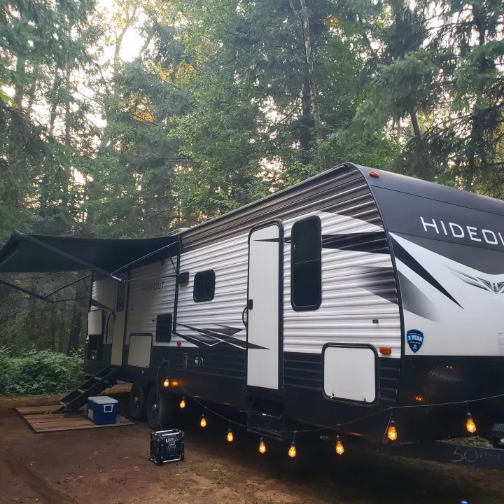 Experience nature with an RV rental! A Hideout travel trailer at a wooded campsite, awning out with string lights.. Keystone RV Hideout Luxury 2021