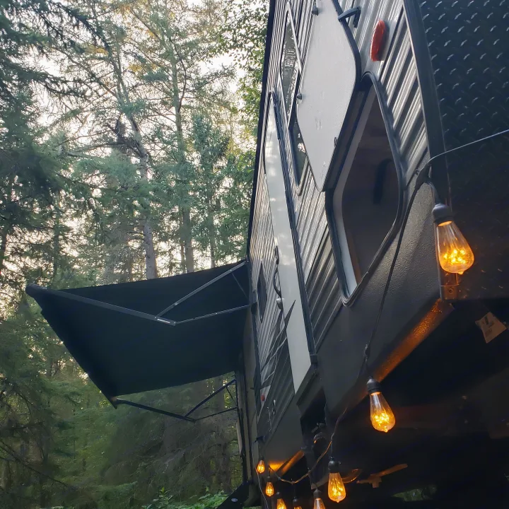 A modern rv rental parked in a tranquil forest, showcasing its extended awning and warm string lights.. Keystone RV Hideout Luxury 2021