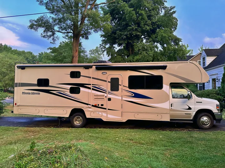 Side view of a beige Winnebago Minnie Winnie RV rental, parked on asphalt with grass. Trees and houses are visible in background.. Winnebago Minnie Winnie 2022