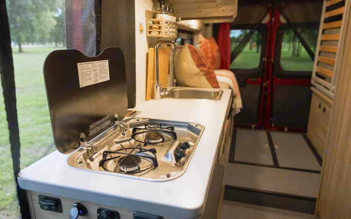 Enjoy an RV rental's compact kitchen with a stove and sink, overlooking nature through a screened window.. Winnebago Solis 2023