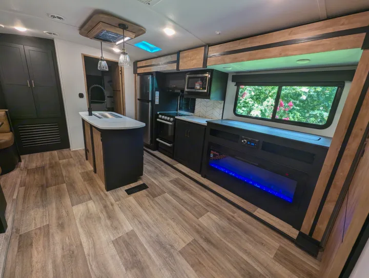 Inside a stylish rv rental: light wood floors, dark cabinets, a kitchen island, and an electric fireplace with blue light.. Keystone RV Outback 2022