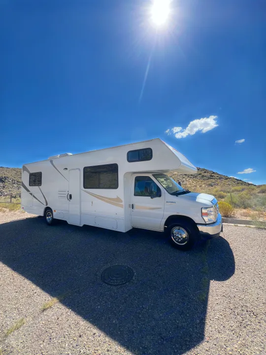 This white RV rental shines under a bright blue sky with sun rays, parked in a desert landscape.. Thor Majestic 2019