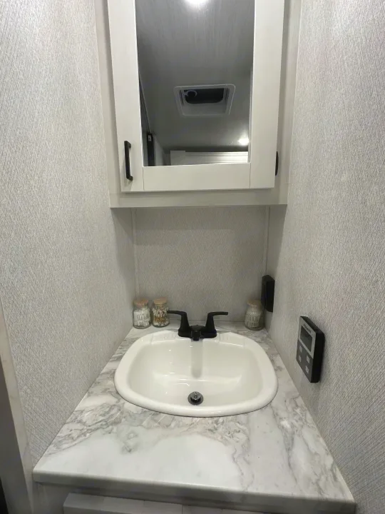 Modern RV rental bathroom vanity with a white sink, black faucet, marble-look counter, and mirrored cabinet.. Entrada East to West 2024