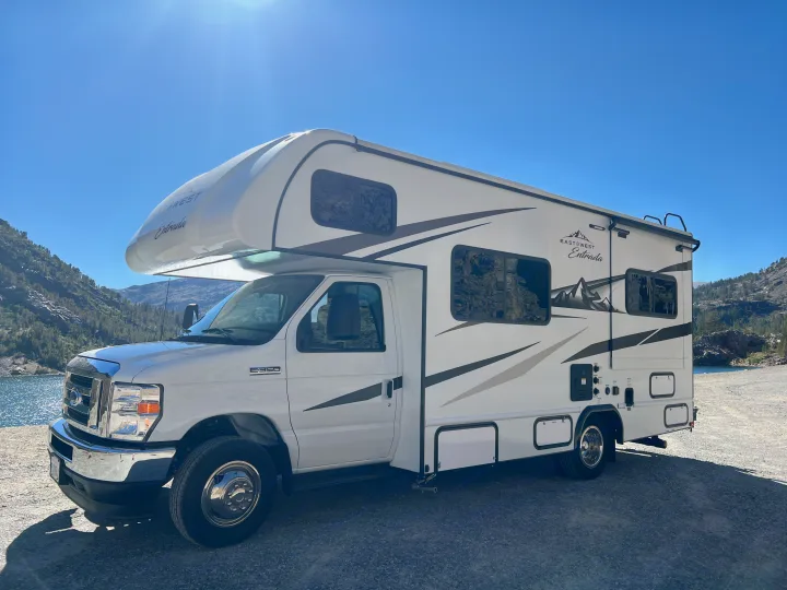 A white EastWest Entrada Class C RV rental is parked by a scenic mountain lake under a bright blue sky.. Entrada East to West 2024