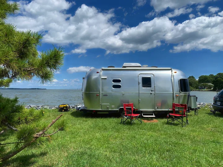 An Airstream RV rental on green grass by a calm bay. Red chairs and a generator are set up for waterfront camping under a cloudy sky.. Airstream Caraval 2021