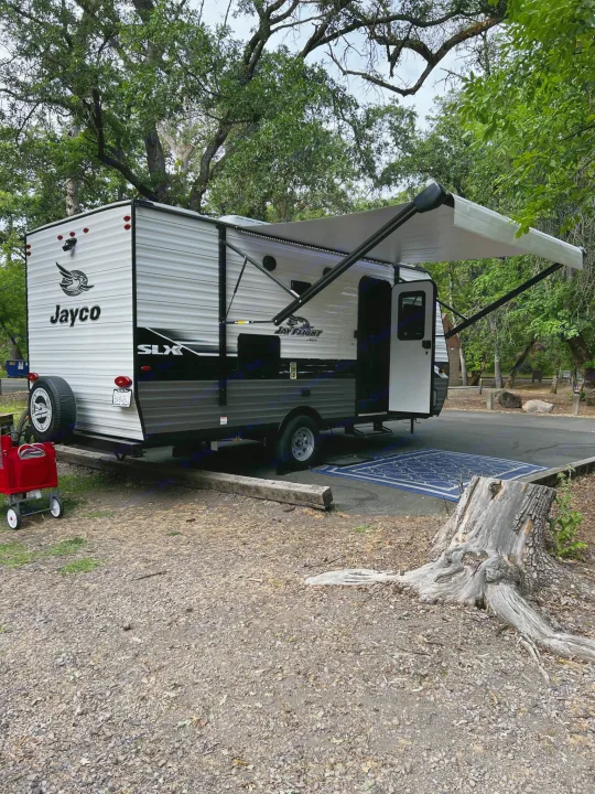 A Jayco rv rental at a peaceful wooded campsite with its awning extended, a blue outdoor rug, and a red wagon nearby.. Jayco Jay Flight SLX 2022