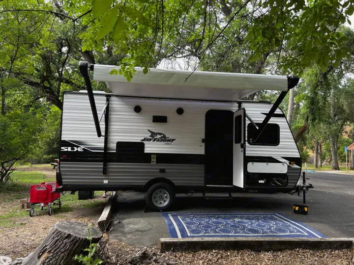 An RV rental, a Jay Flight travel trailer, is set up at a leafy campsite with an open awning and blue outdoor rug.. Jayco Jay Flight SLX 2022