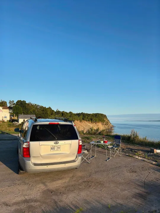 Perfect RV rental getaway spot! Silver minivan, folding chairs, and table face a serene blue sea with cliffs. Quebec plate: N86 ZPF.. KIA SEDONNA 2008