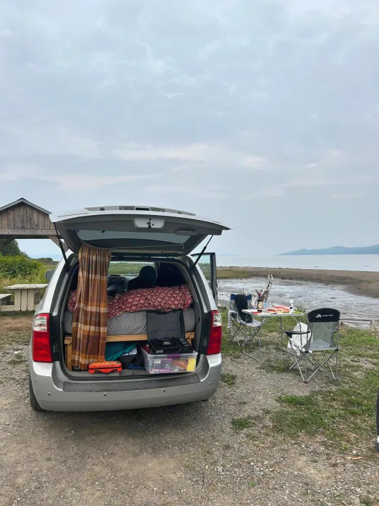 Silver minivan set up like an RV rental, parked by a bay. The back is open showing a bed; chairs & table outside.. KIA SEDONNA 2008