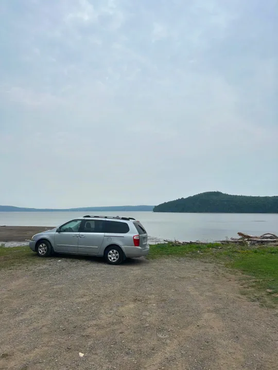 Silver minivan by scenic lake with forested hill under cloudy sky. Perfect for an RV rental-style adventure.. KIA SEDONNA 2008