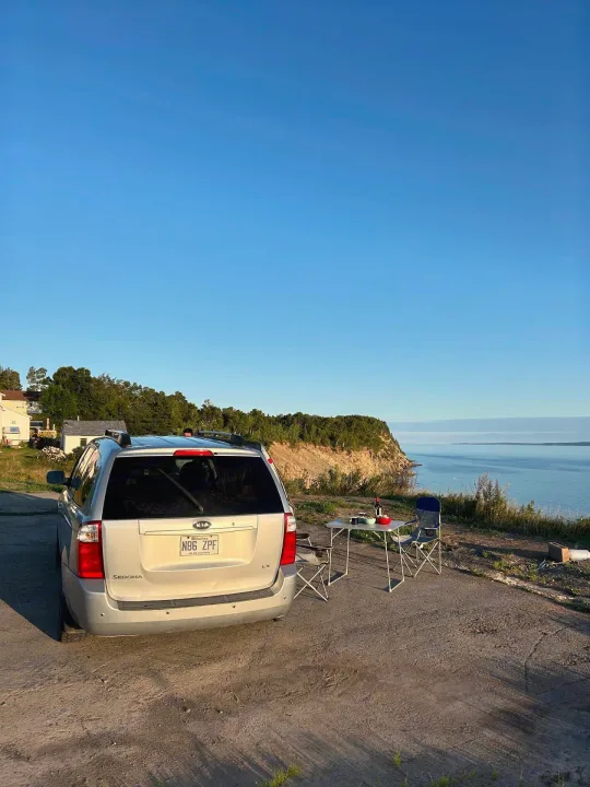 Silver Kia Sedona minivan parked overlooking the ocean from a cliff. Camping table & chairs suggest an RV rental-like adventure.. KIA SEDONNA 2008
