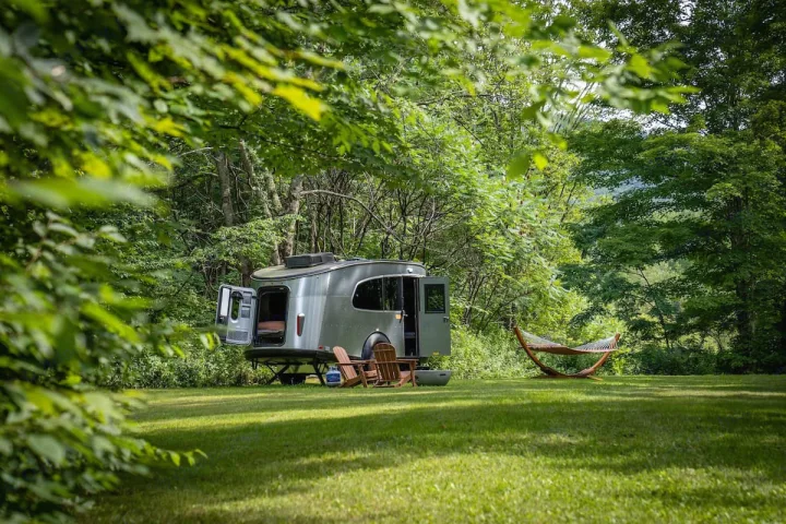 Discover peace with an RV rental! A modern silver travel trailer, chairs, and hammock await on a lush green lawn near a forest.. Airstream Base Camp 2023
