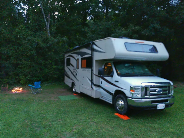 An RV rental is parked at a grassy campsite during dusk. A warm campfire glows, with a blue folding chair beside it.. Coachmen Leprechaun 230 CB 2018