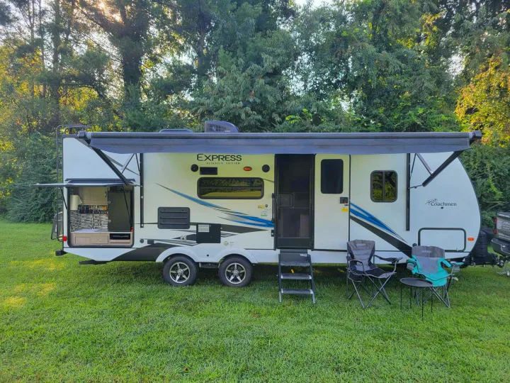 This white RV rental, with an extended awning, outdoor kitchen, and camping chairs, is set on a green lawn with trees in the background.. Coachmen Freedom Express 248RBS 2020