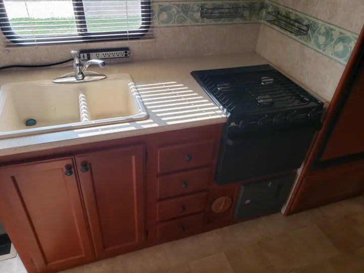RV rental kitchen: double sink, gas stove, power outlets, and wood cabinets. Sunlight streams through window blinds.. Four Winds Freedom Elite 2010