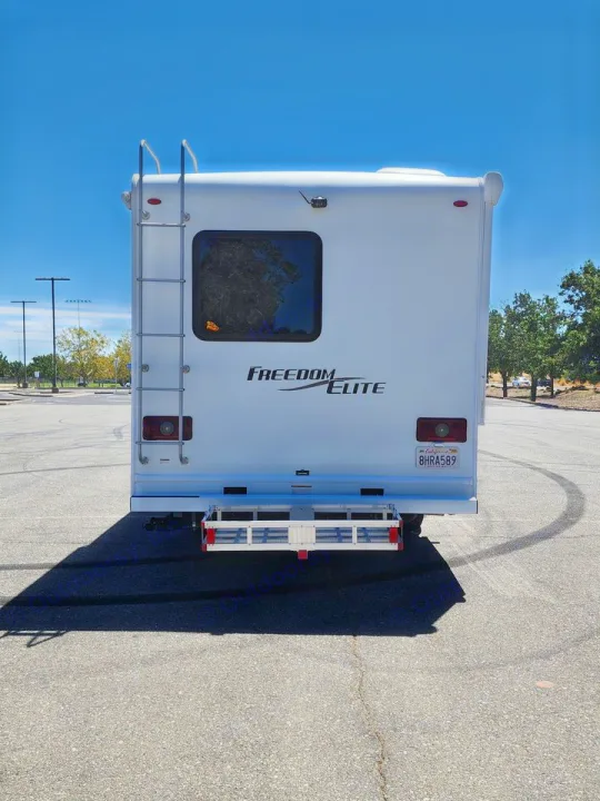 Rear view of a white **RV rental**, featuring a ladder, "FREEDOM ELITE" branding, and a cargo carrier in a sunny parking lot.. Thor Freedom Elite 2019