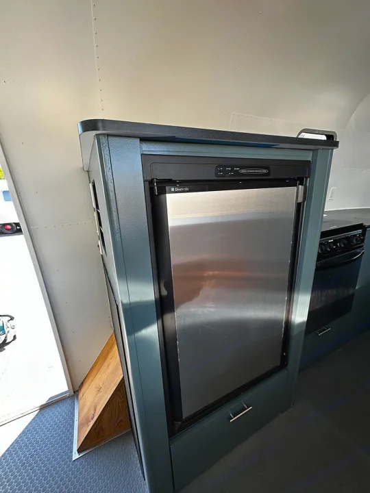 Inside an RV rental kitchen, featuring a Dometic refrigerator, dark countertop, and teal cabinets, with a ramp at the door.. Airstream Custom 2006