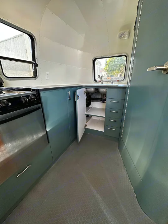 This RV rental kitchen features stylish teal cabinets, a stove, and a sink. The open cabinet reveals plumbing.. Airstream Custom 2006