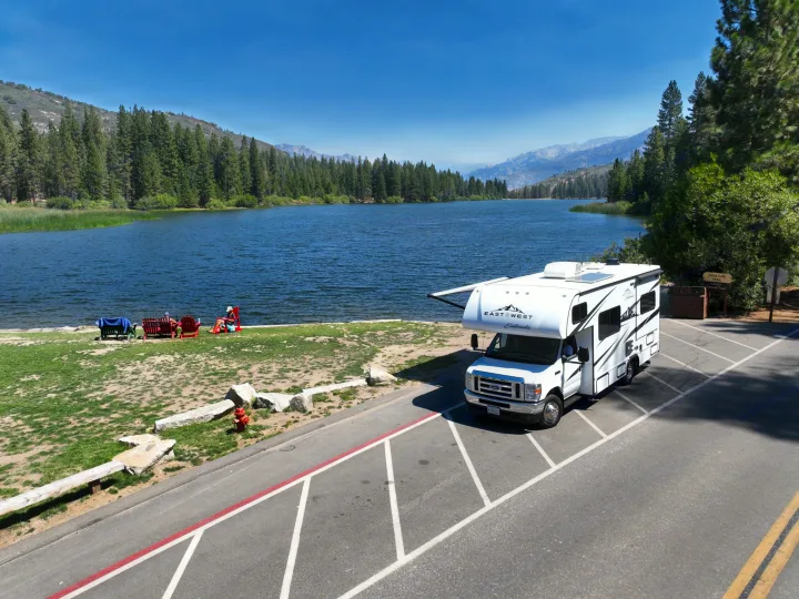 White RV rental parked by a tranquil lake, framed by pine trees and mountains. People relax on the shore enjoying the sunny outdoor scene.. Entrada East to West 2024