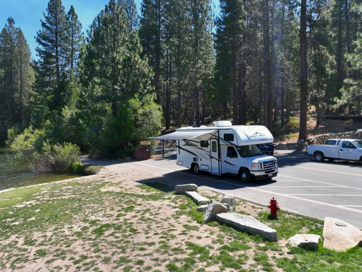 A white RV rental with an extended awning is parked by Hume Lake, surrounded by a scenic pine forest.. Entrada East to West 2024