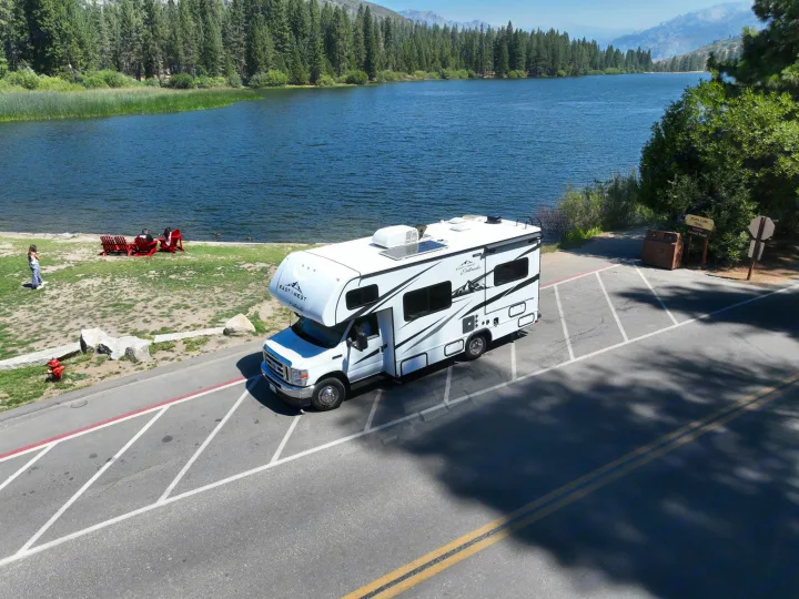 An rv rental, a white East West Entrada RV, is parked by the shores of a scenic lake, surrounded by green forests and mountains.. Entrada East to West 2024