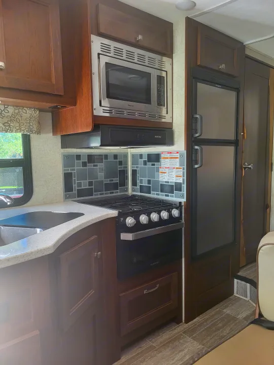 RV rental kitchen featuring stainless appliances: microwave, stove, fridge, and sink. Includes dark wood cabinets & tile backsplash.. Forester Forester Motorhome 2020