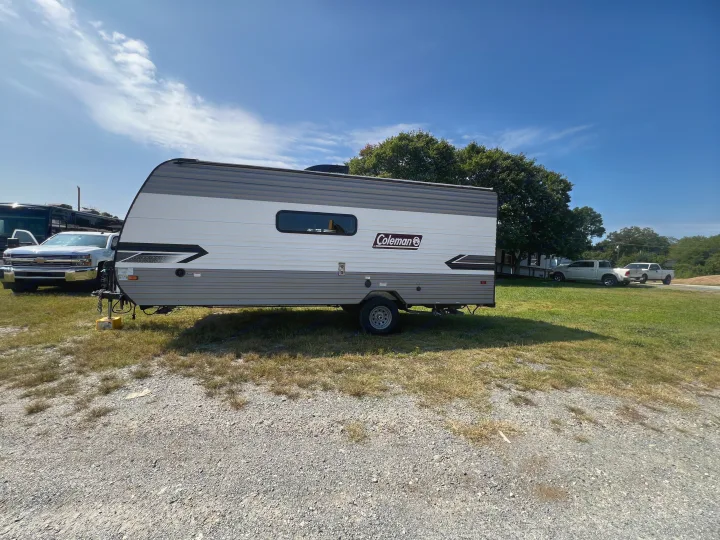 A white and grey Coleman travel trailer, ideal for an rv rental, parked on a mixed gravel and grass lot on a sunny day.. Coleman Lantern 18 2024