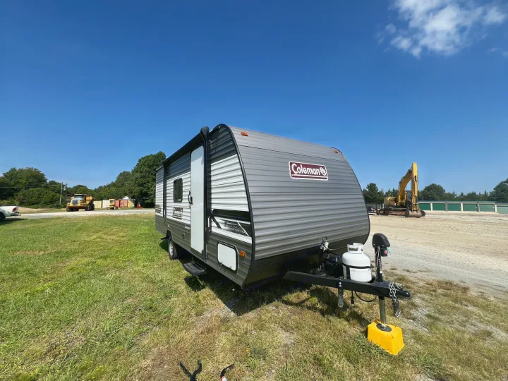Coleman travel trailer RV rental, grey, parked on grass. Clear blue sky, excavator & storage units in background.. Coleman Lantern 18 2024