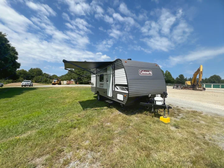 Sunny day shows an RV rental Coleman travel trailer with awning, parked on grass near construction equipment under a blue sky.. Coleman Lantern 18 2024