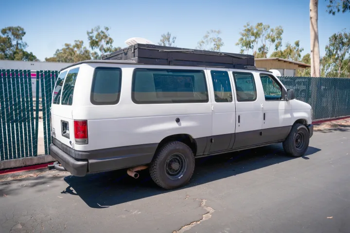 A white Ford van, converted for travel with a roof rack and rugged tires, an ideal RV rental for your next adventure.. Ford Econoline 2008
