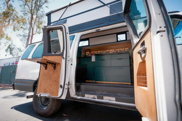 This RV rental features an open side door revealing a stylish mobile kitchen, pop-top, and rear door table. Perfect for van life adventures.. Ford Econoline 2008