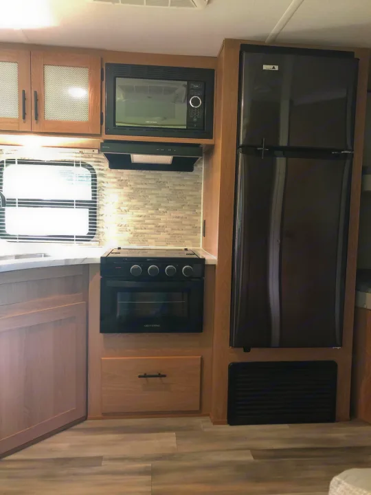 Bright RV rental kitchen with modern black appliances (microwave, stove, fridge), light wood cabinets & sleek backsplash.. Forest River Prime Time Avenger 24BHS 2023