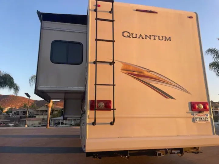 Rear of a Quantum motorhome RV rental with ladder, extended slide-out, and California plate by a lake.. Thor Quantum 2019