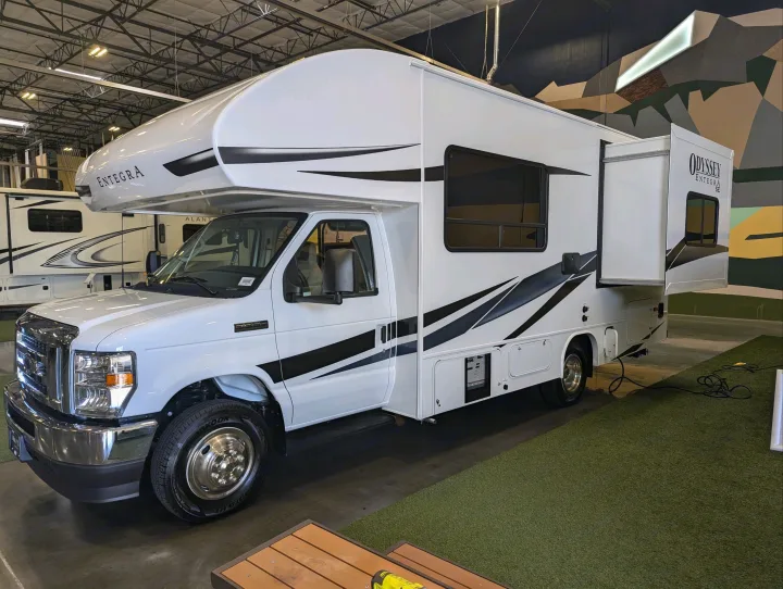 A sleek white Entegra Odyssey Class C RV with black decals, a prime option for rv rental, on display indoors.. Entegra Odyssey SE 2025