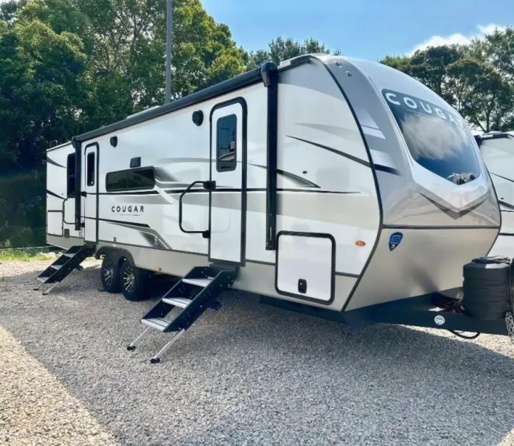 Modern gray and white Cougar travel trailer, ideal for an rv rental, parked on gravel with steps extended under sunny skies.. Keystone RV Cougar 2024