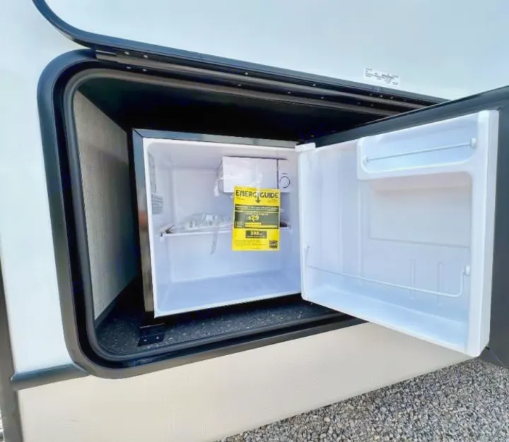 RV rental: An exterior RV compartment is open, revealing a white mini-fridge with its door ajar and an energy guide label.. Keystone RV Cougar 2024