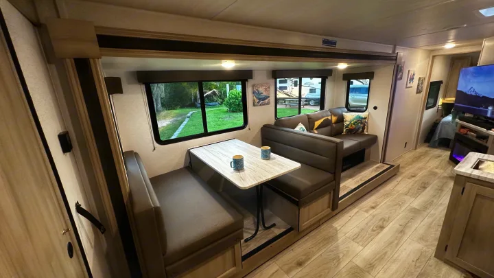 Cozy RV rental interior featuring a dinette with a table and a comfortable sofa, seen through large windows overlooking nature.. East to West Silver Lake 3250QB 2025