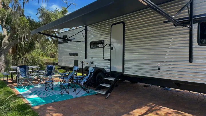 This RV rental campsite features an extended awning, outdoor seating on a mat, and a picnic table by the water, perfect for relaxation.. East to West Silver Lake 3250QB 2025