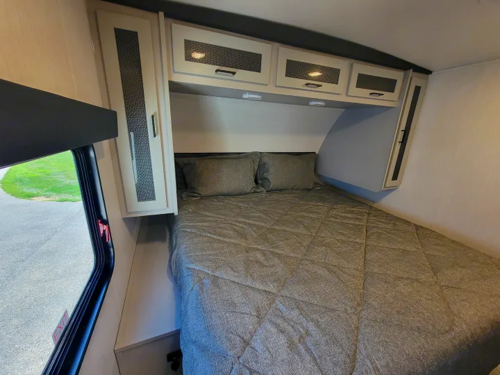 Cozy RV rental bedroom with a comfy grey bed, modern overhead cabinets, and a window offering outdoor views.. Heartland RVs North Trail 2023