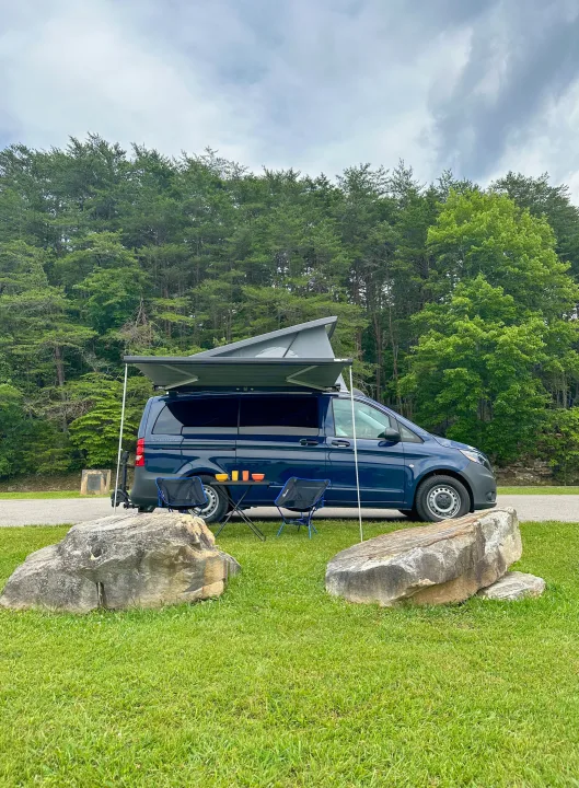 A blue camper van with pop-top & awning, like an RV rental, is set up with chairs & table on grass by a forest.. Mercedes-Benz Metris 2023