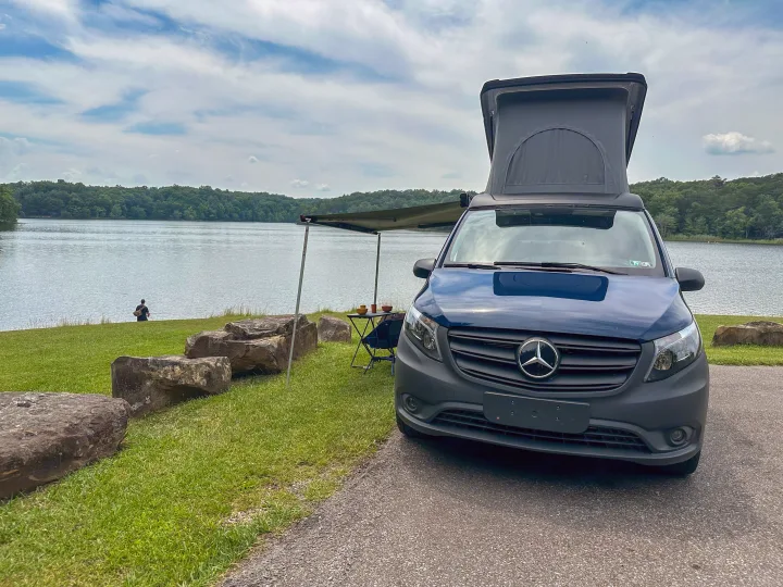 Scenic rv rental: Mercedes pop-top camper van with awning by a tranquil lake, surrounded by green forests, perfect for nature getaways.. Mercedes-Benz Metris 2023