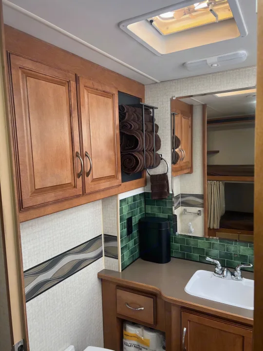 Cozy RV rental bathroom with wood cabinets, green tile backsplash, and sink, reflecting bunk beds beyond.. Winnebago Minnie Winnie 2017