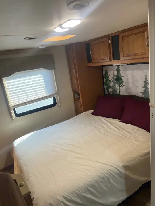 Cozy RV rental bedroom featuring a comfortable bed, window with blinds, overhead cabinets, and a nature-themed headboard.. Winnebago Minnie Winnie 2017