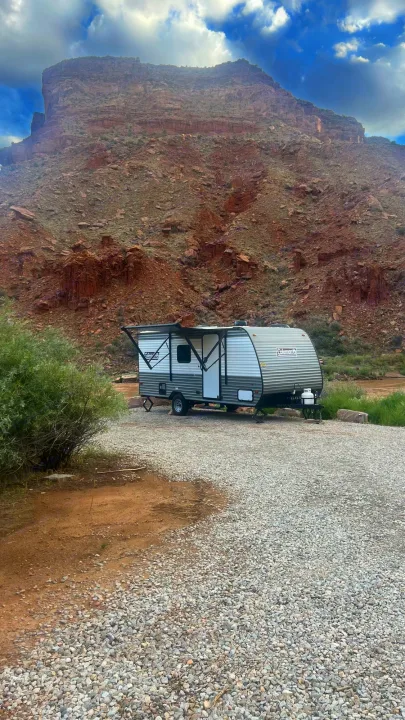 Scenic RV rental camping: travel trailer parked on gravel, with an awning, red rock mountains & river in background.. Dutchmen Coleman Lantern LT 2024