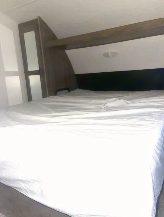 This RV rental bedroom features a clean bed with white sheets, a dark headboard, and overhead storage.. Pioneer Other 2023
