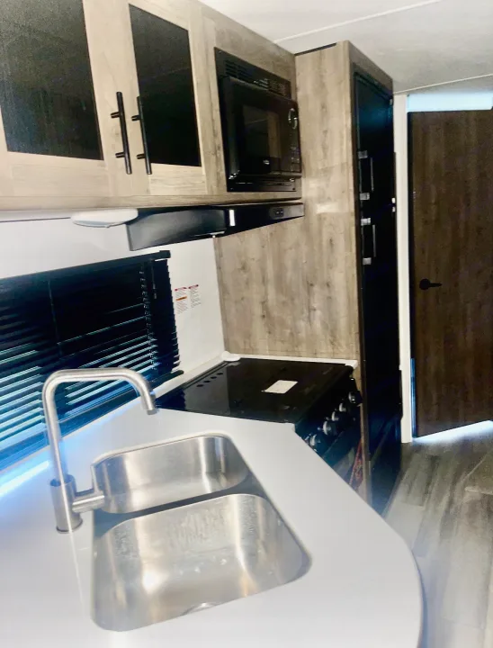 Modern RV rental kitchen features a stainless steel sink, stove, microwave, and light wood cabinets for compact living.. Pioneer Other 2023