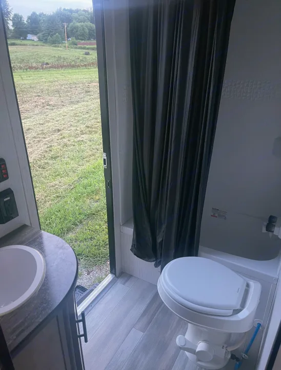 Inside an RV rental: a compact bathroom with toilet, sink, and shower, overlooking a green field through the open door.. Pioneer Other 2023