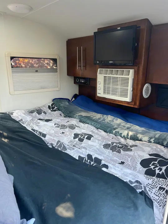 This RV rental's compact sleeping area features a bed with patterned bedding, a window, mounted TV, and AC unit.. nuCamp T@G 2018