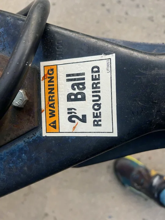 Close-up of an RV rental trailer hitch showing a "WARNING 2" Ball REQUIRED" sticker on dark metal for safe towing.. nuCamp T@G 2018