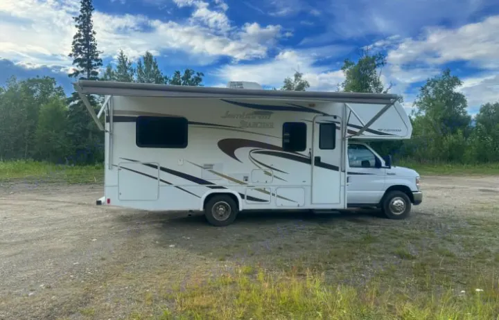 White Class C RV rental parked on a gravel lot with its awning extended. Surrounded by trees under a cloudy sky, ideal for trips.. Jamboree Searcher 2014